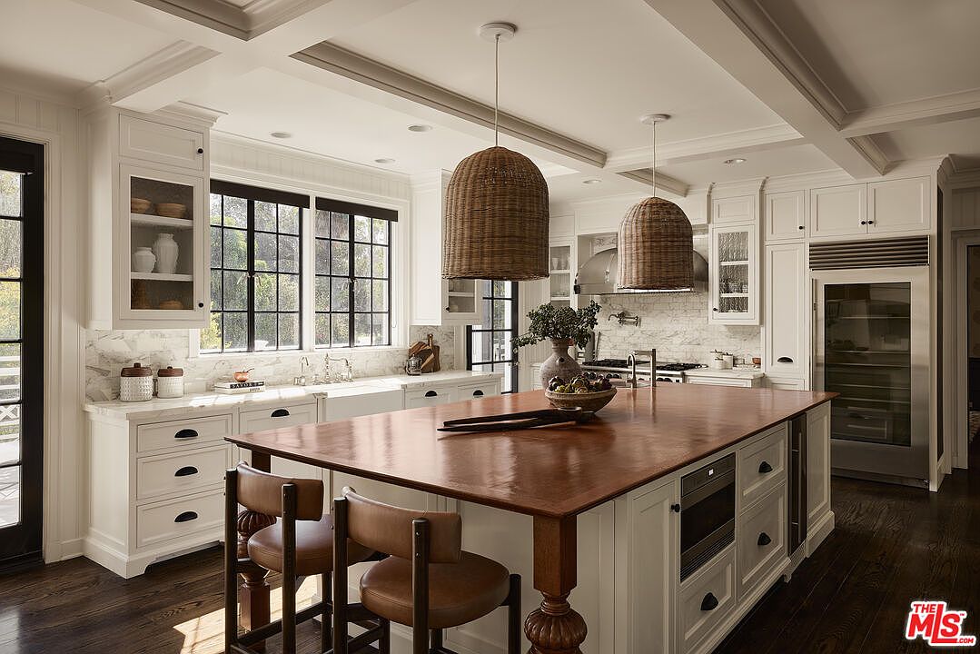 This is a bright and spacious kitchen featuring custom white cabinetry, a large wooden island with seating, and high-end appliances. The kitchen is well-lit with natural light from the large windows and pendant lighting above the island. The dark wood flooring adds warmth and contrast to the space, creating an inviting and luxurious atmosphere.