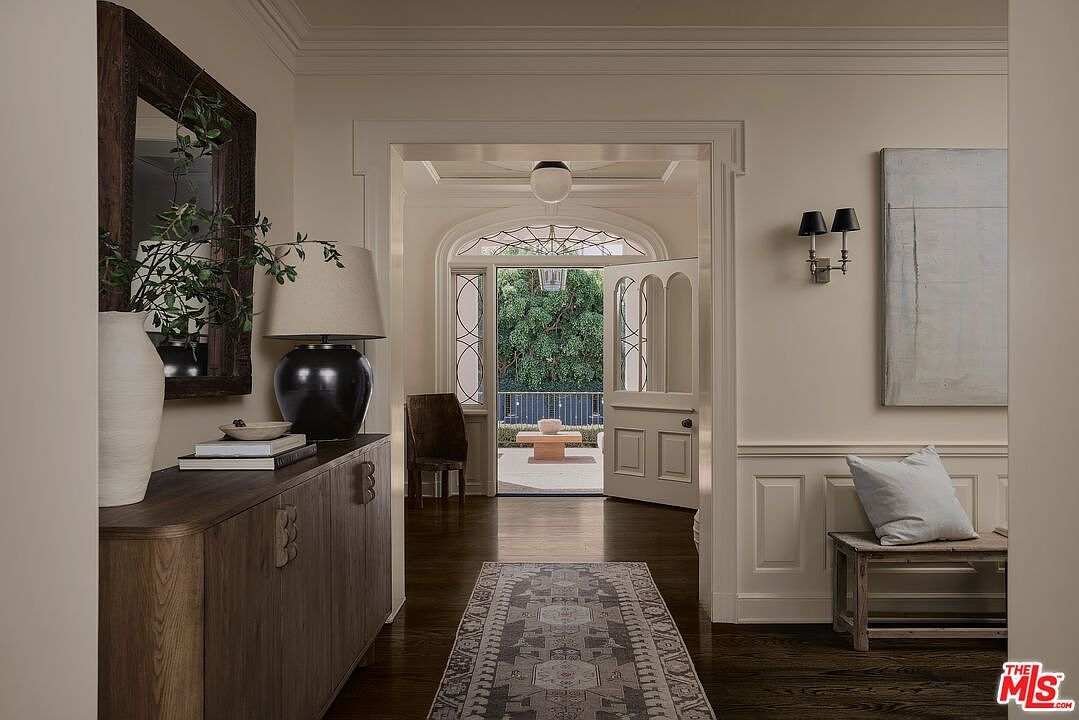 This interior shot showcases a well-lit hallway with dark hardwood floors and neutral-toned walls. A wooden console table with a lamp and decorative items sits on the left, while a bench with a pillow is on the right. The hallway leads to an open doorway, providing a glimpse of an outdoor patio area, creating a sense of depth and connection to the outdoors.