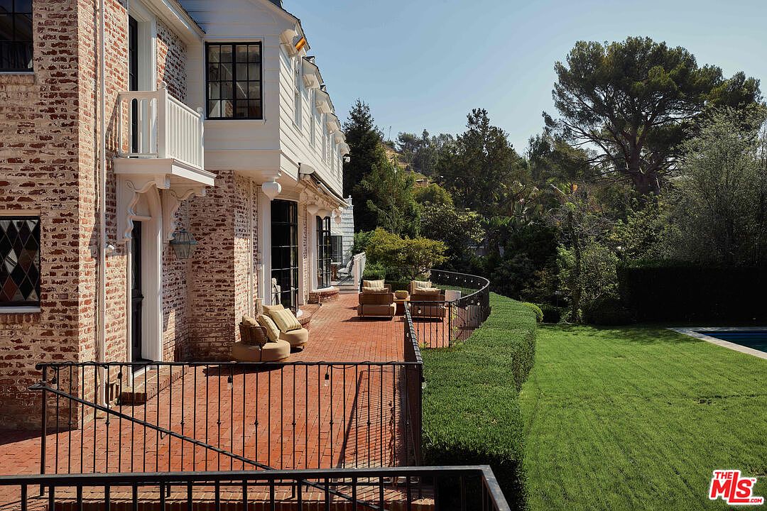 This image showcases a charming patio/deck area of a house, featuring a brick-paved surface and a wrought-iron railing. Comfortable outdoor seating is arranged on the deck, suggesting a relaxing space for entertaining or enjoying the surrounding greenery. The house itself has a classic architectural style with brick and white accents.
