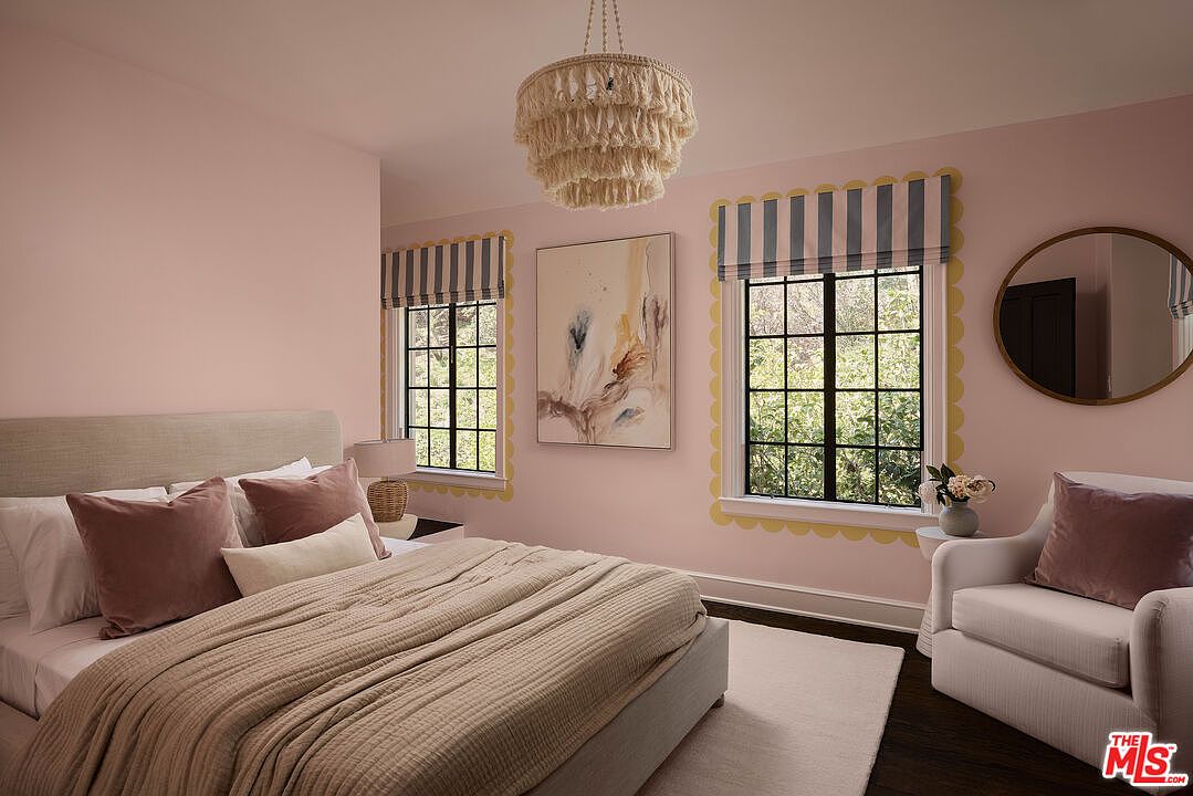 This is a charming bedroom featuring a bed with plush pillows and a cozy blanket, complemented by a stylish chandelier and framed artwork. The room is painted in a soft pink hue, with windows framed by striped valances and decorative trim, adding a touch of whimsy. An armchair and round mirror complete the inviting and comfortable space.