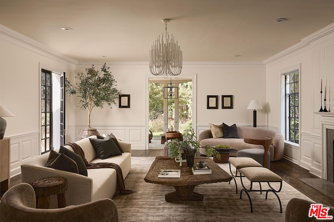 This is an interior shot of a living room featuring two sofas, a large coffee table, and a chandelier. The room is decorated in neutral tones with dark wood floors and white trim, creating a sophisticated and inviting atmosphere. The open doorway leads to an outdoor area, enhancing the sense of space and connection to nature.