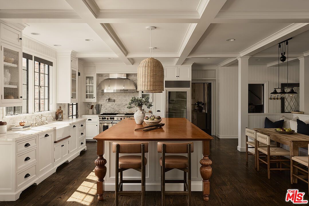This is a bright and spacious kitchen featuring white cabinetry, marble countertops, and stainless steel appliances. A large wooden island with seating anchors the space, while a woven pendant light adds a touch of texture. The dark hardwood floors provide a beautiful contrast to the light-colored elements, creating a warm and inviting atmosphere.