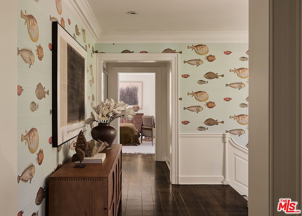 This interior shot showcases a hallway adorned with whimsical fish-themed wallpaper, complemented by dark hardwood flooring. A wooden console table with decorative objects sits against the wall, while an open doorway leads to another room, creating depth and visual interest. The space feels stylish and inviting, with a unique design element that adds character.
