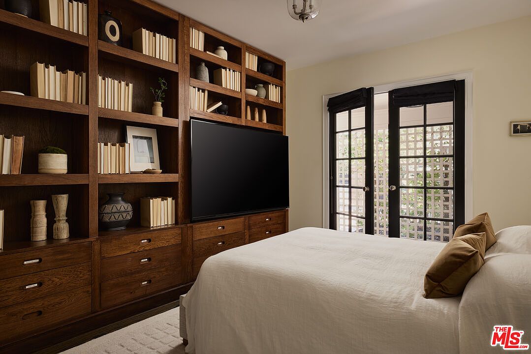 This is a primary bedroom featuring a built-in wooden bookcase and entertainment center with a large television. The room has a neutral color palette with a large bed dressed in white linens and brown pillows. French doors with black frames lead to an outdoor area, providing natural light.
