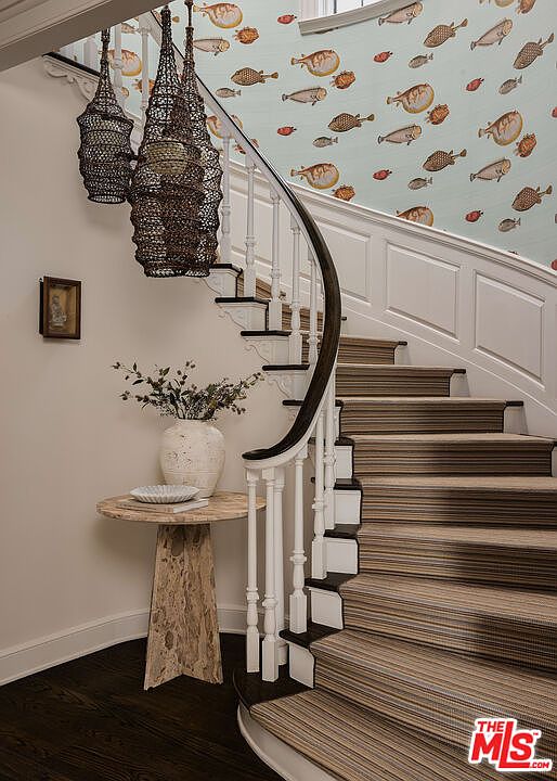 This interior shot showcases a beautifully designed hallway and staircase. The staircase features striped carpeting, white railings with a dark wood handrail, and black accents on the steps. A round table with a vase of flowers sits at the base of the stairs, and unique woven light fixtures hang above, adding a touch of elegance and character to the space.
