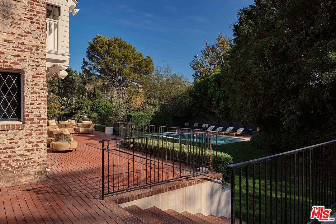 This image showcases a charming outdoor patio area, featuring a brick-paved surface and comfortable seating arrangements. A black iron railing provides a safety barrier and adds a touch of elegance. In the background, a well-maintained pool area with lounge chairs offers a relaxing retreat, all surrounded by lush greenery and mature trees.