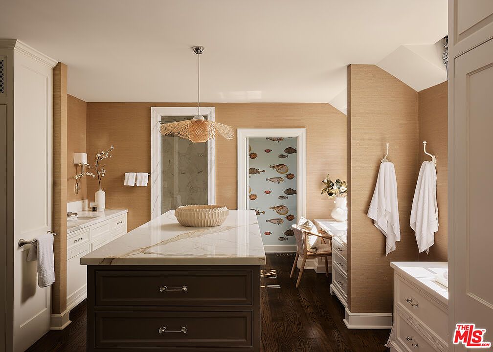 This is a well-appointed primary bathroom featuring a central island with a marble countertop and dark wood cabinetry. The walls are covered in a textured wallpaper, and there's a doorway leading to a room with fish-themed wallpaper. The bathroom also includes a vanity area with white cabinetry and a separate area with towel hooks, creating a luxurious and functional space.