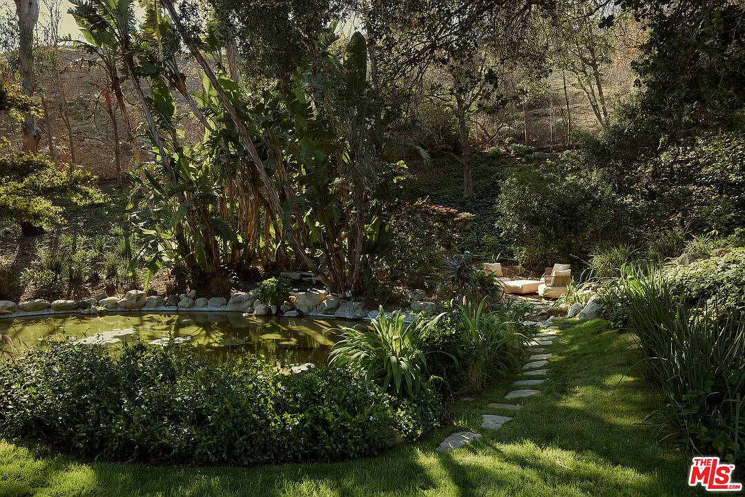 This image showcases a lush and serene garden featuring a tranquil pond surrounded by rocks and various plants. A stone pathway leads to a seating area with lounge chairs, creating a peaceful retreat. The overall impression is one of natural beauty and relaxation, perfect for enjoying the outdoors.