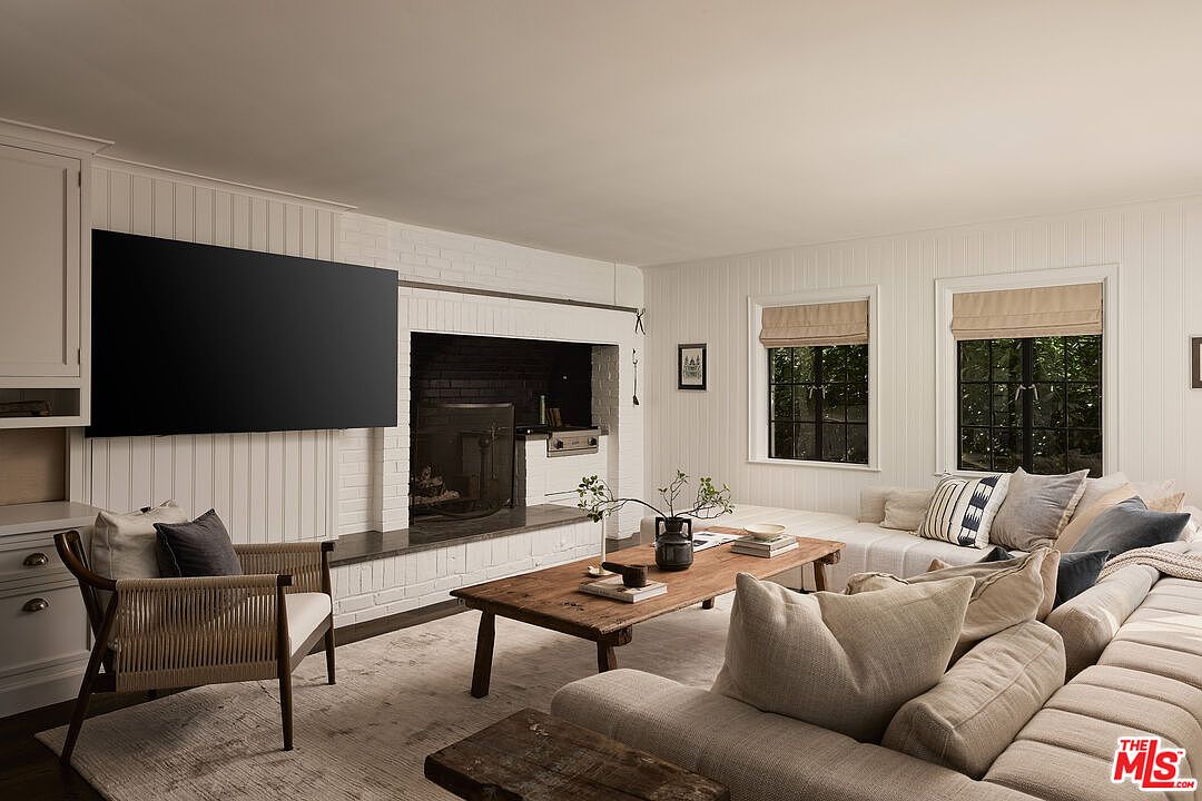The living room features a large flat-screen TV mounted above a white brick fireplace. A comfortable sectional sofa is arranged around a rustic wooden coffee table, creating a cozy and inviting atmosphere. Natural light filters through the windows, enhancing the room's warm and neutral color palette.
