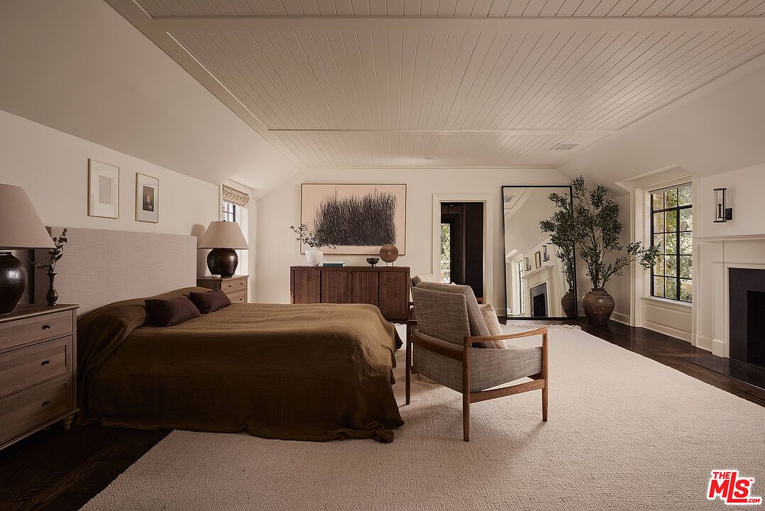 This primary bedroom features a neutral color palette with a large bed, wooden nightstands, and a dresser with artwork above. A comfortable armchair sits on a large area rug, and a full-length mirror reflects the fireplace and window. The room exudes a calm and sophisticated atmosphere.