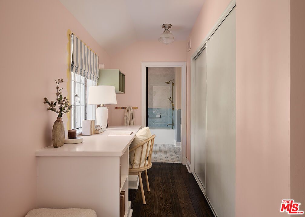 This interior shot showcases a cozy office space with a built-in white desk, a light wood chair, and a window with striped valances. The room is painted in a soft pink hue, complemented by dark wood flooring and a glimpse into an adjacent bathroom. The overall impression is a blend of functionality and understated elegance.