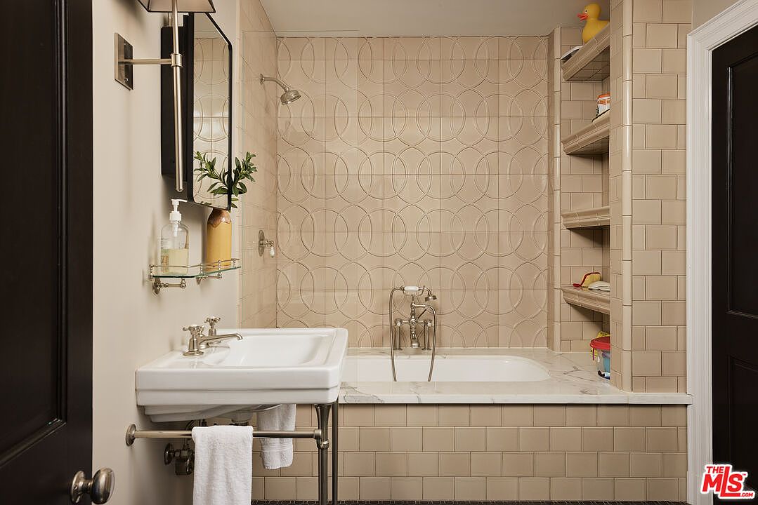 This bathroom features a classic design with beige tiled walls and a unique circular pattern above the bathtub. A pedestal sink with chrome fixtures and a glass shelf adds to the vintage charm. Built-in shelving to the right of the tub provides storage, and the overall impression is clean and well-maintained.