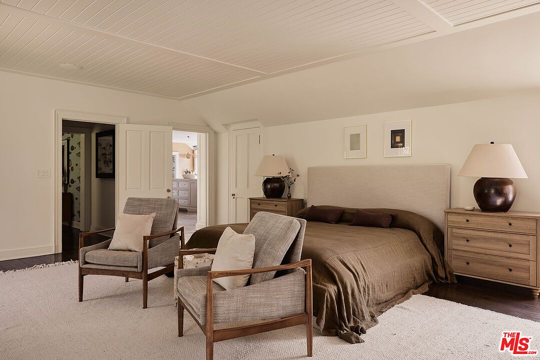 This is a well-lit primary bedroom featuring a neutral color palette. The room includes a bed with a light beige headboard and brown bedding, two matching wooden nightstands with lamps, and two armchairs with light-colored cushions. The room has a calm and inviting atmosphere, enhanced by the natural light and the clean, uncluttered design.