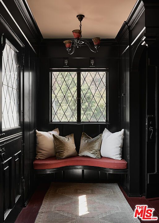 This interior shot showcases a hallway or entryway with dark, paneled walls and a built-in bench beneath a window. The window features a diamond-patterned design, and the bench is adorned with pillows. A vintage chandelier hangs from the ceiling, adding to the room's unique character and charm.
