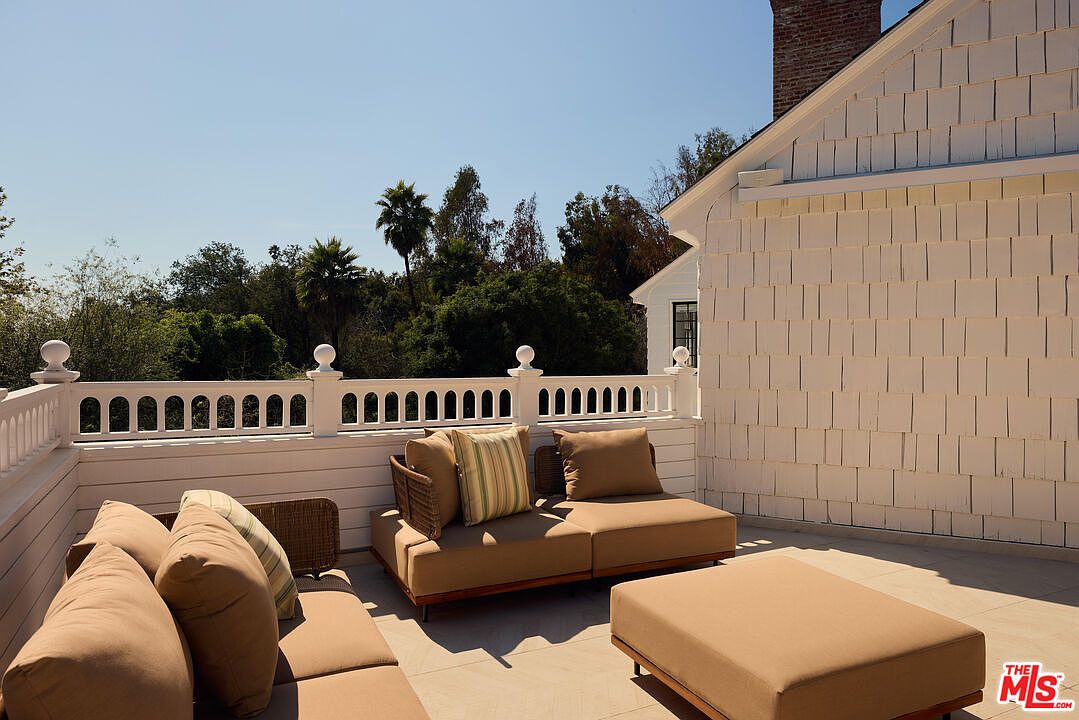 This image showcases a beautifully designed outdoor patio or deck area. The space features comfortable lounge furniture with neutral-toned cushions, complemented by a white railing and architectural details on the adjacent building. Lush greenery in the background adds a sense of privacy and tranquility, making it an ideal space for relaxation and outdoor entertaining.