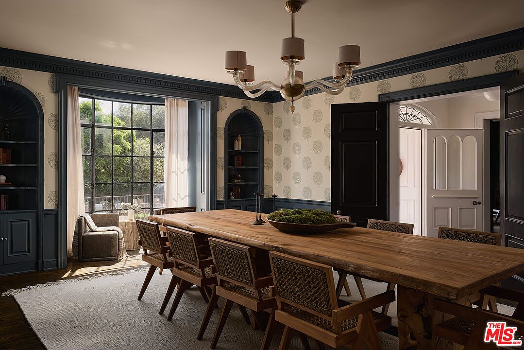 This is an interior shot of a dining room featuring a large wooden table with woven chairs. The room is decorated with a patterned wallpaper and dark blue trim, complemented by a modern chandelier. Natural light floods the space through a large window, creating a warm and inviting atmosphere.