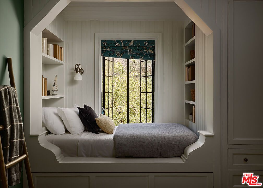 This image showcases a cozy window seat in a bedroom, featuring built-in bookshelves and a comfortable bed with pillows and a blanket. The window offers a view of lush greenery, and the overall design creates a relaxing and inviting atmosphere. The room is well-lit and appears to be part of a thoughtfully designed home.