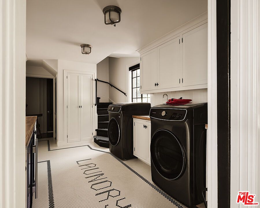 This is a well-designed laundry room featuring a black front-load washer and dryer set, white cabinetry, and a unique tiled floor with the word "LAUNDRY" spelled out. The room is bright and clean, with modern fixtures and a practical layout, suggesting a functional and stylish space for laundry tasks. The perspective is from the doorway, giving a full view of the room's layout.