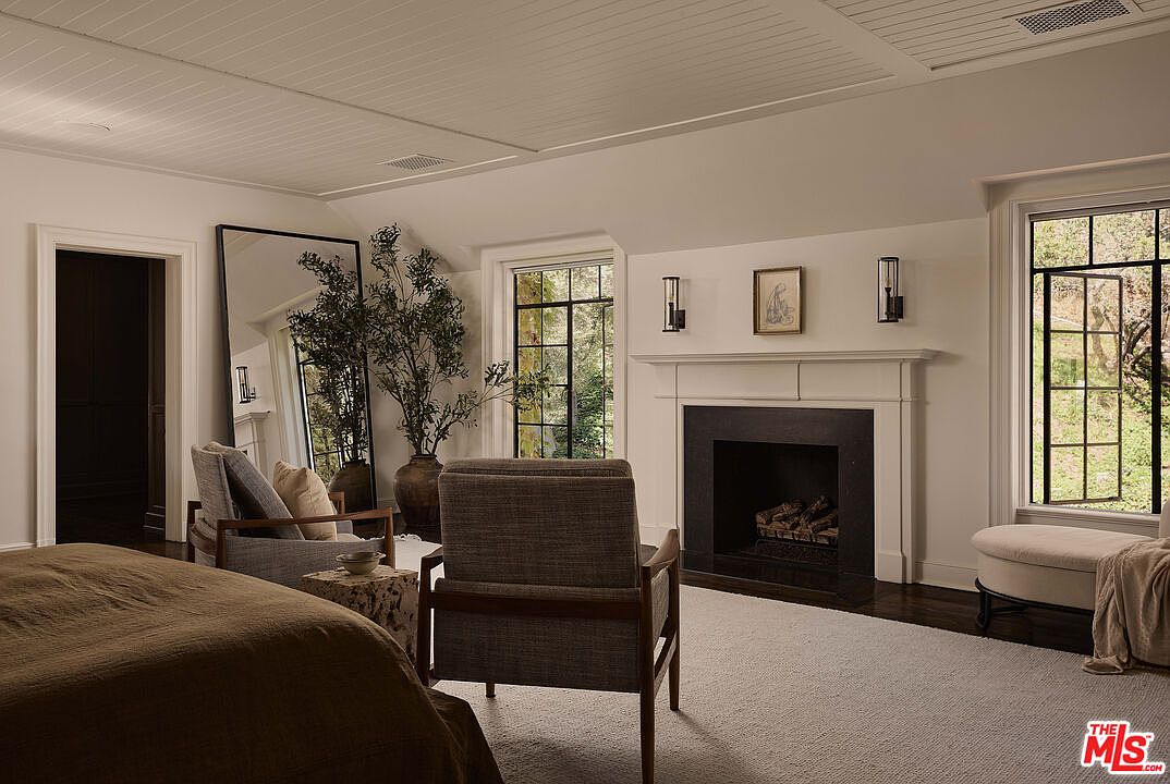 This is a cozy primary bedroom featuring a fireplace, two armchairs, and a large mirror. The room has a neutral color palette with white walls and a light-colored carpet. Natural light streams in through the windows, creating a warm and inviting atmosphere.