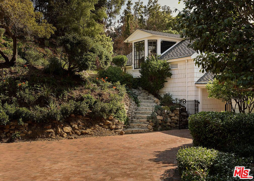 This image showcases the front exterior of a charming home, featuring a brick driveway leading up to the house. Stone steps ascend a landscaped hillside to the side of the house, adding visual interest and character. The house itself has a white exterior with a distinctive window feature on the upper level, complemented by lush greenery and mature trees.