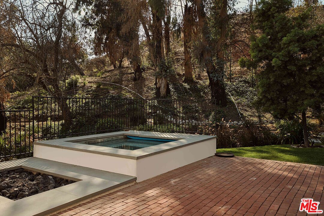This image showcases an outdoor spa area with a modern aesthetic. The spa is surrounded by a brick patio and a fire pit filled with dark rocks. A black metal fence separates the spa area from a natural, wooded hillside, creating a private and serene setting.