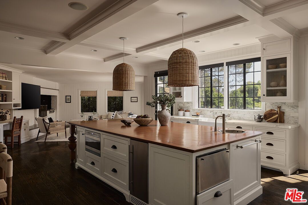 This is a bright and spacious kitchen featuring a large center island with a wooden countertop and white cabinetry. The kitchen is well-lit with natural light from the large windows and complemented by stylish pendant lighting. The dark hardwood floors add a touch of elegance to the space, creating a warm and inviting atmosphere.