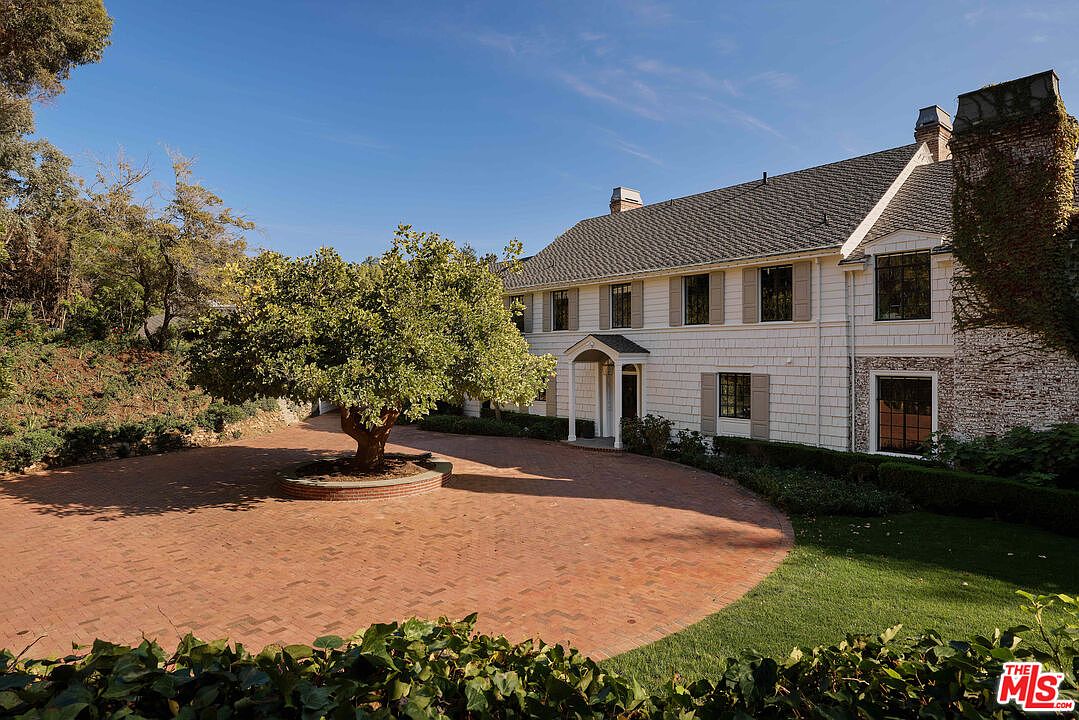 This is a front exterior view of a large, two-story house with a brick driveway and a mature tree in the center. The house features white siding, dark-framed windows with light shutters, and a covered entryway. A brick chimney covered in ivy adds character to the right side of the house, and manicured landscaping surrounds the property, enhancing its curb appeal.