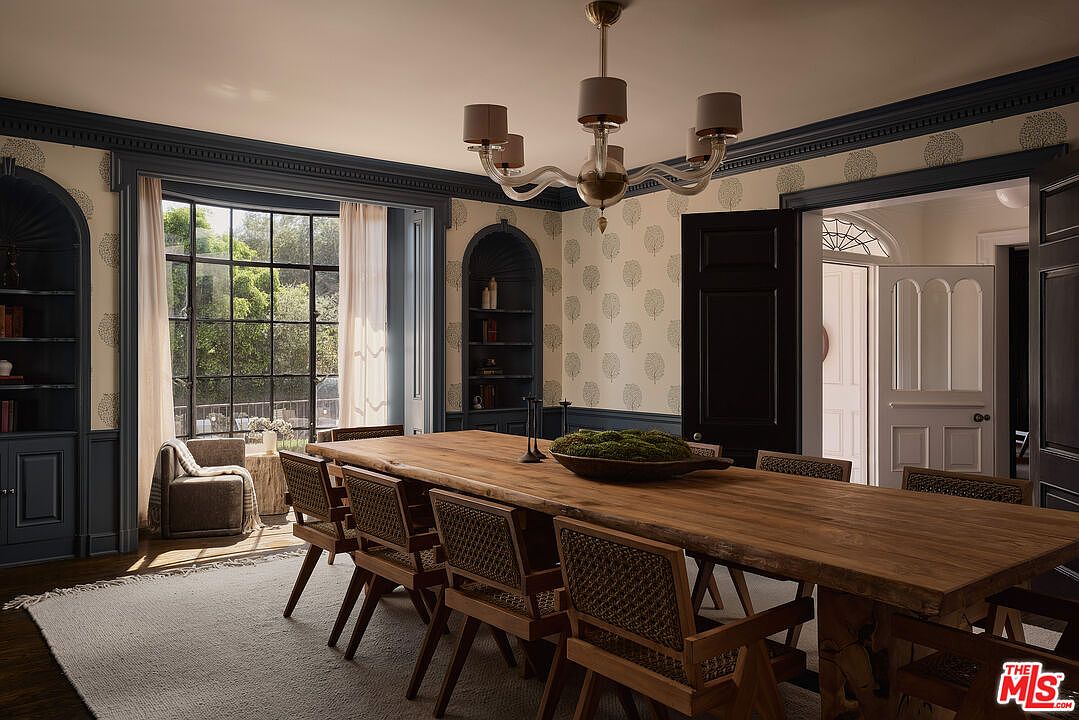 This is an interior shot of a dining room featuring a long, rustic wooden table with woven chairs. The room is decorated with patterned wallpaper and dark blue trim, complemented by a modern chandelier. Natural light streams in through a large window, creating a warm and inviting atmosphere.
