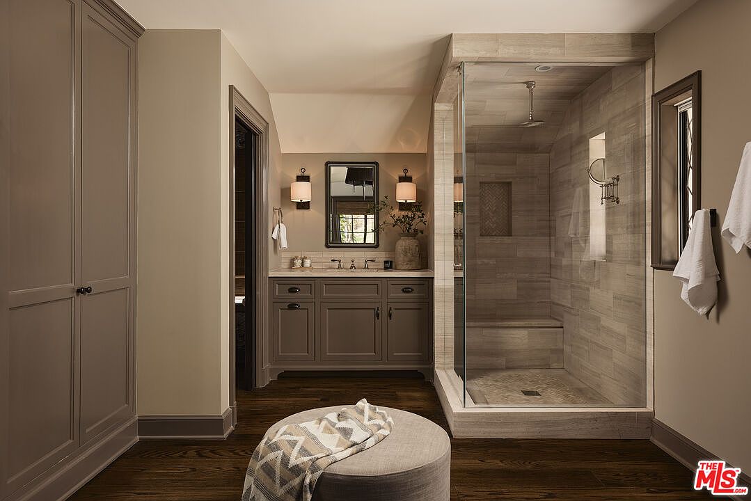 This is a well-appointed primary bathroom featuring a double vanity with a marble countertop, dark wood cabinetry, and sconce lighting. A glass-enclosed shower with horizontal tile and a built-in bench adds a luxurious touch. The dark wood flooring and neutral wall colors create a warm and inviting atmosphere.