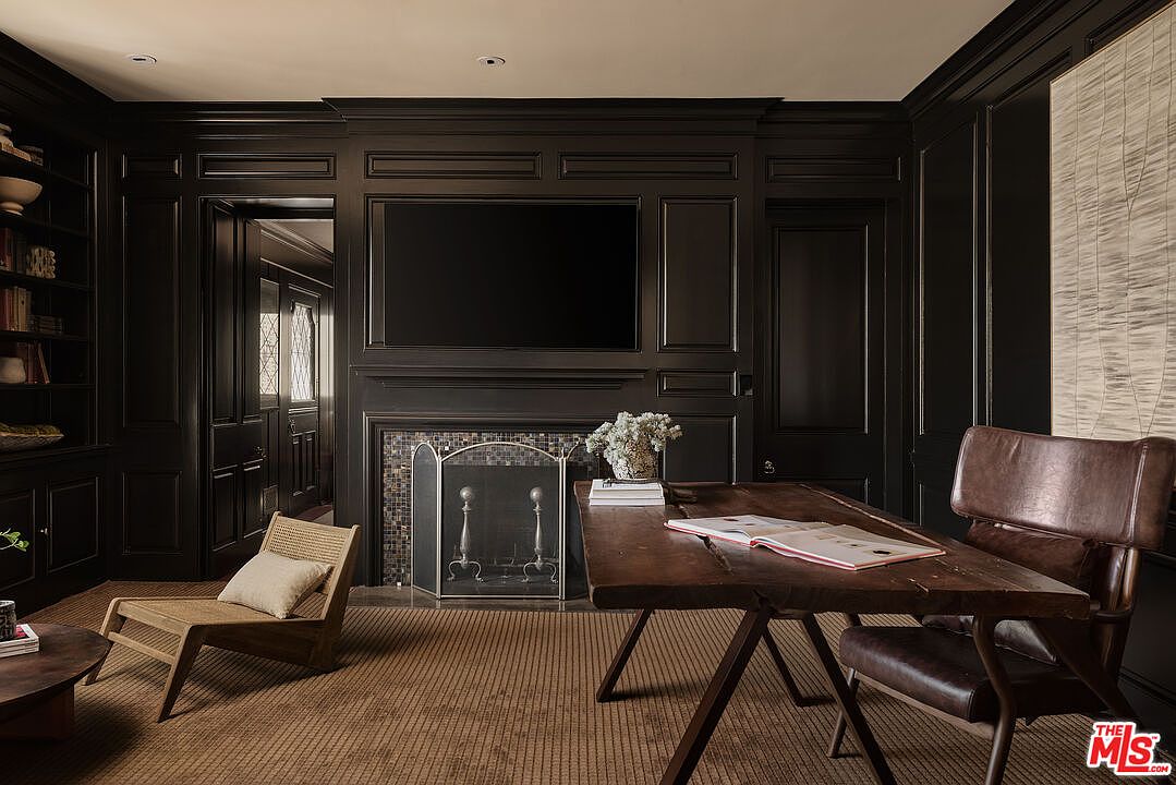 This is an interior shot of a sophisticated home office or study, featuring dark wood paneling and built-in shelving. A large flat-screen television is mounted above the fireplace, and a rustic wooden desk with a leather chair sits in the foreground. The room exudes a sense of classic elegance and professional comfort.
