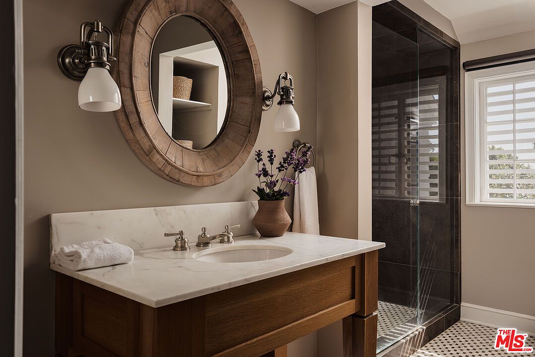 This bathroom features a rustic-chic design with a wooden vanity topped with a marble countertop and a round wooden-framed mirror. The walls are painted in a neutral tone, complemented by bronze sconces flanking the mirror. A glass-enclosed shower with dark tiling and a window with blinds complete the space, creating a warm and inviting atmosphere.