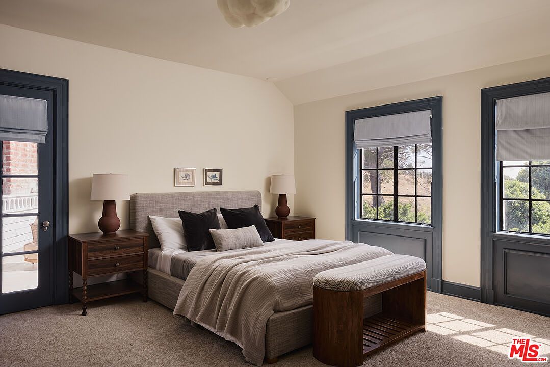This is a primary bedroom featuring a neutral color palette with blue accents on the door and window frames. The room is furnished with a bed, two nightstands with lamps, and a bench at the foot of the bed. Natural light streams in through the windows, creating a serene and inviting atmosphere.