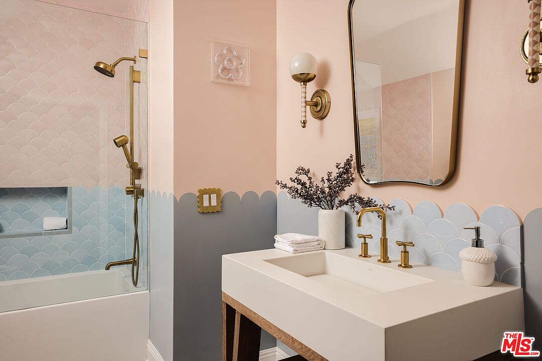 This is a stylish bathroom featuring a modern sink with a gold faucet, a decorative mirror, and unique wall tiling in pastel colors. The walls are painted in a two-tone scheme with a scalloped edge separating the colors, adding a playful touch. The overall design creates a bright and cheerful atmosphere.