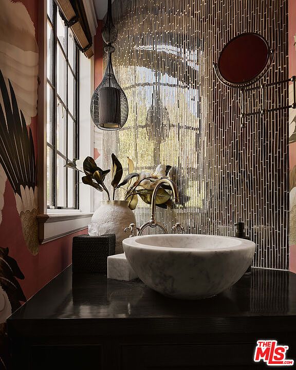 This is a stylish bathroom featuring a vessel sink made of marble, complemented by a modern faucet and a unique vase with decorative plants. The wall behind the sink is adorned with a textured, vertical tile design, and a round mirror is mounted on the wall. The overall aesthetic is sophisticated and artistic, creating a luxurious and inviting space.