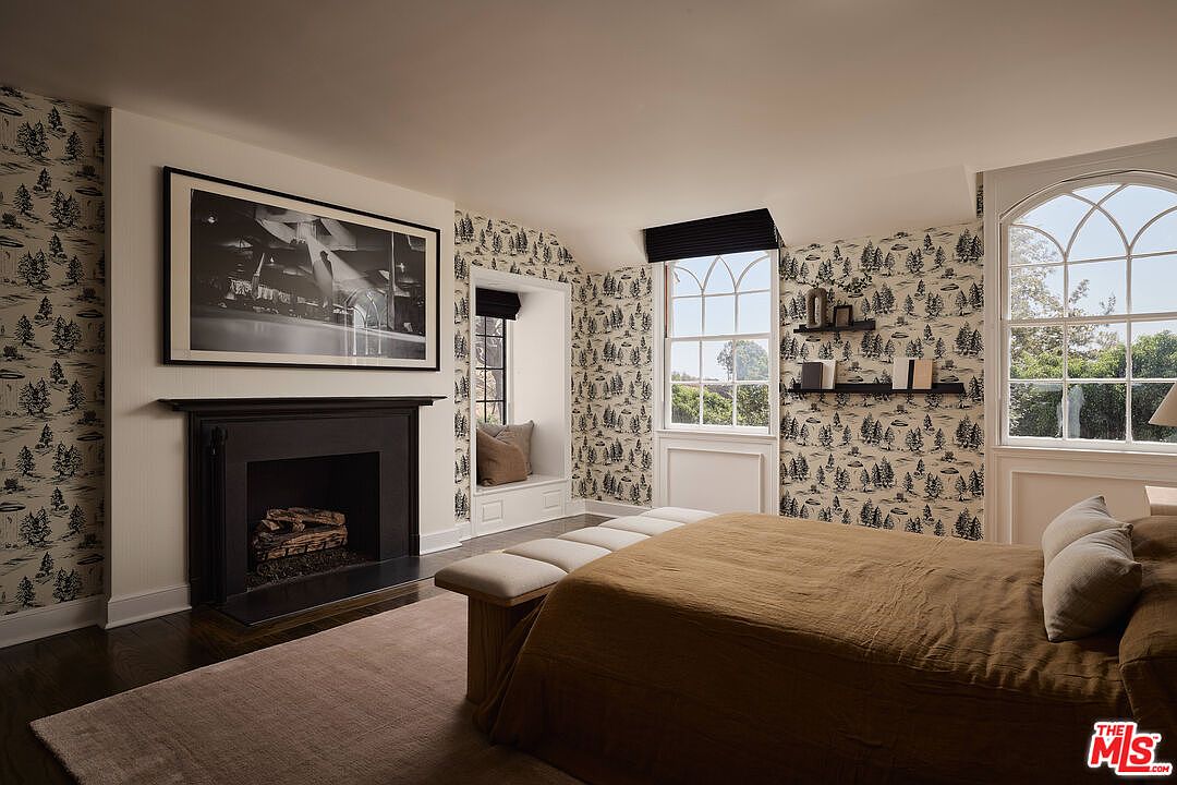 This is a primary bedroom featuring patterned wallpaper, a dark wood fireplace with a large black and white photograph above it, and two windows providing natural light. The room is furnished with a bed covered in a brown duvet and a bench at the foot of the bed, creating a cozy and inviting atmosphere. The dark wood floors and neutral rug add warmth to the space.
