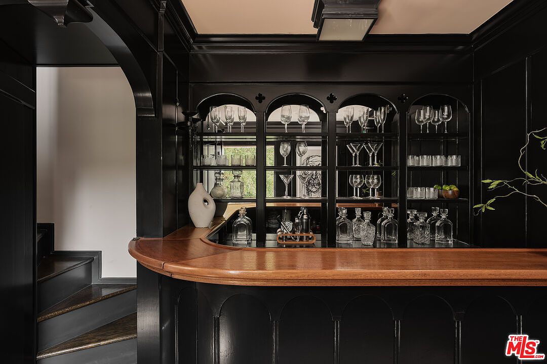 This image showcases a sophisticated home bar area, featuring dark cabinetry with arched detailing and a warm wood countertop. The shelves are stocked with glassware and bottles, creating an inviting atmosphere for entertaining. The space is well-lit, highlighting the textures and finishes of the bar.