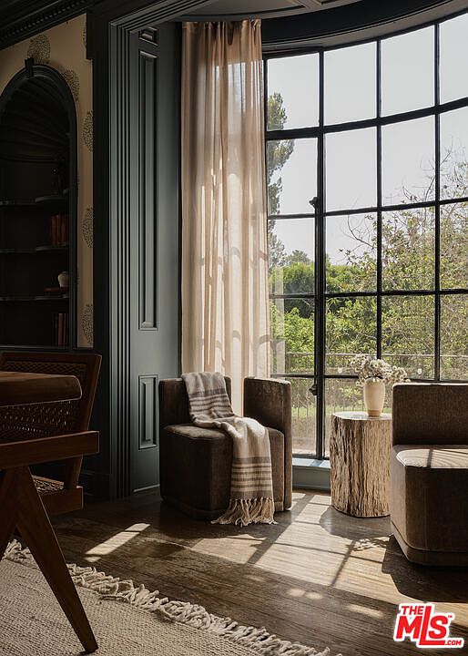 This interior shot showcases a cozy living room area with a large window providing ample natural light. The room features comfortable seating, including armchairs with a throw blanket, and a unique wooden side table with flowers. The dark trim and neutral color palette create a sophisticated and inviting atmosphere.