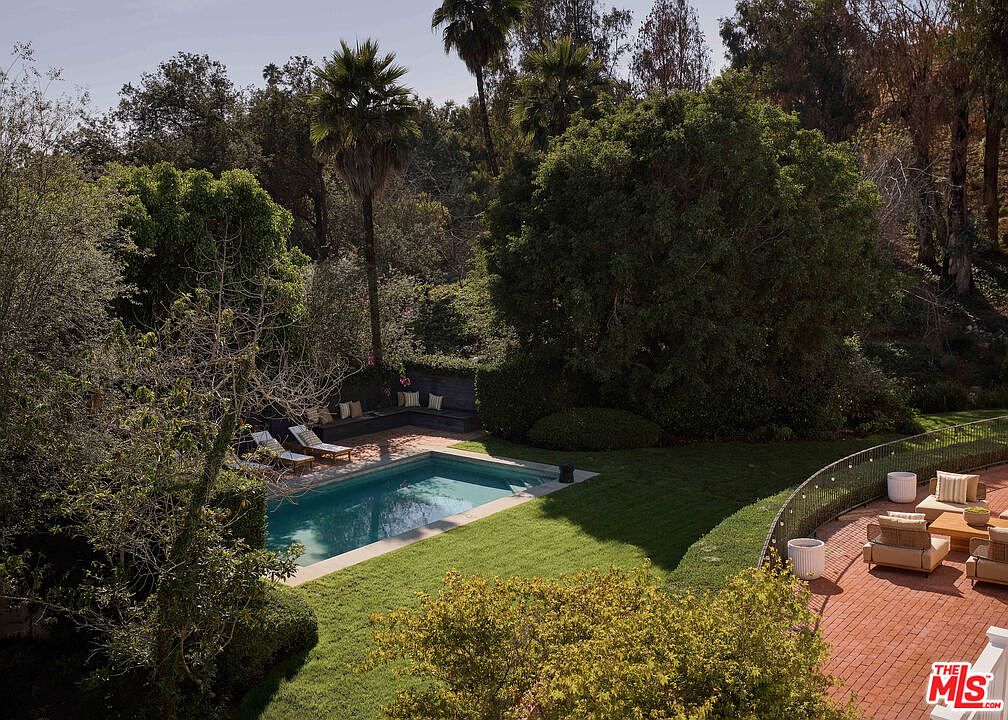 This image showcases a luxurious backyard pool area, complete with a rectangular pool surrounded by lush green grass and mature trees. Lounge chairs are positioned near the pool, and a brick patio with outdoor seating is visible in the background, suggesting a relaxing and private outdoor living space. The scene evokes a sense of tranquility and upscale living.