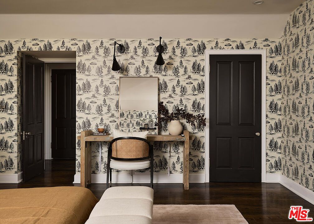 The image showcases a stylish bedroom interior featuring a unique wallpaper with a forest and UFO design. A wooden desk with a mirror and decorative items sits against the wall, complemented by a chair and a bench. The room has dark wood flooring and is illuminated by sconces, creating a modern yet whimsical atmosphere.