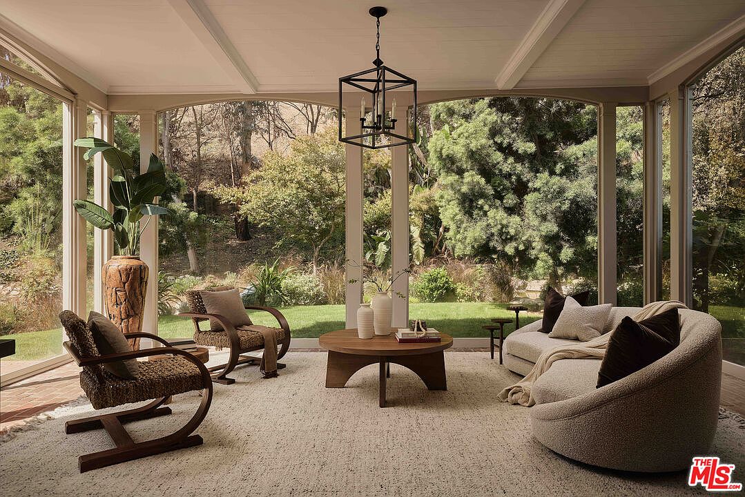 This sunroom features large windows offering views of the surrounding greenery, creating a seamless connection with nature. The room is furnished with comfortable seating, including chairs and a curved sofa, arranged around a round coffee table. A modern chandelier hangs from the ceiling, adding a touch of elegance to the space.