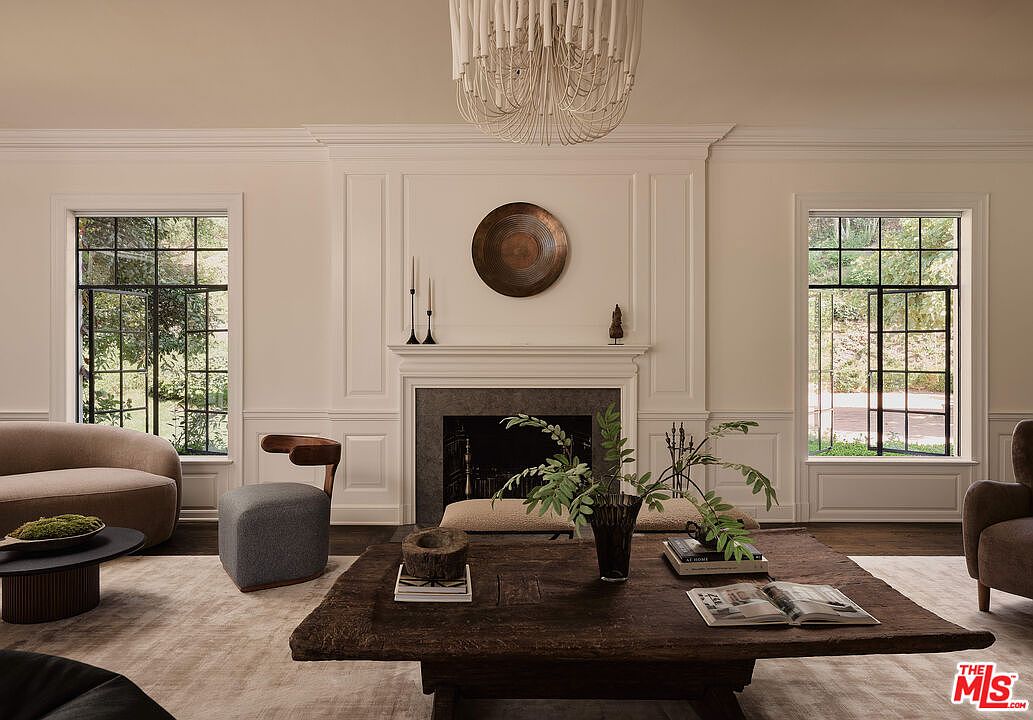 This is an interior shot of a living room featuring a fireplace with a decorative plate above it, flanked by two large windows that let in natural light. The room is furnished with a dark wood coffee table, a curved sofa, and a gray chair, all set on a light-colored rug. The overall aesthetic is elegant and comfortable, with a focus on natural materials and a neutral color palette.