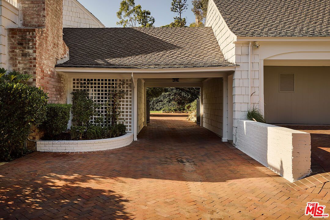 This image showcases a charming garage exterior with a brick-paved driveway leading through a covered carport. The structure features a combination of brick and white siding, complemented by a trellis with greenery. The overall impression is one of classic elegance and well-maintained property.