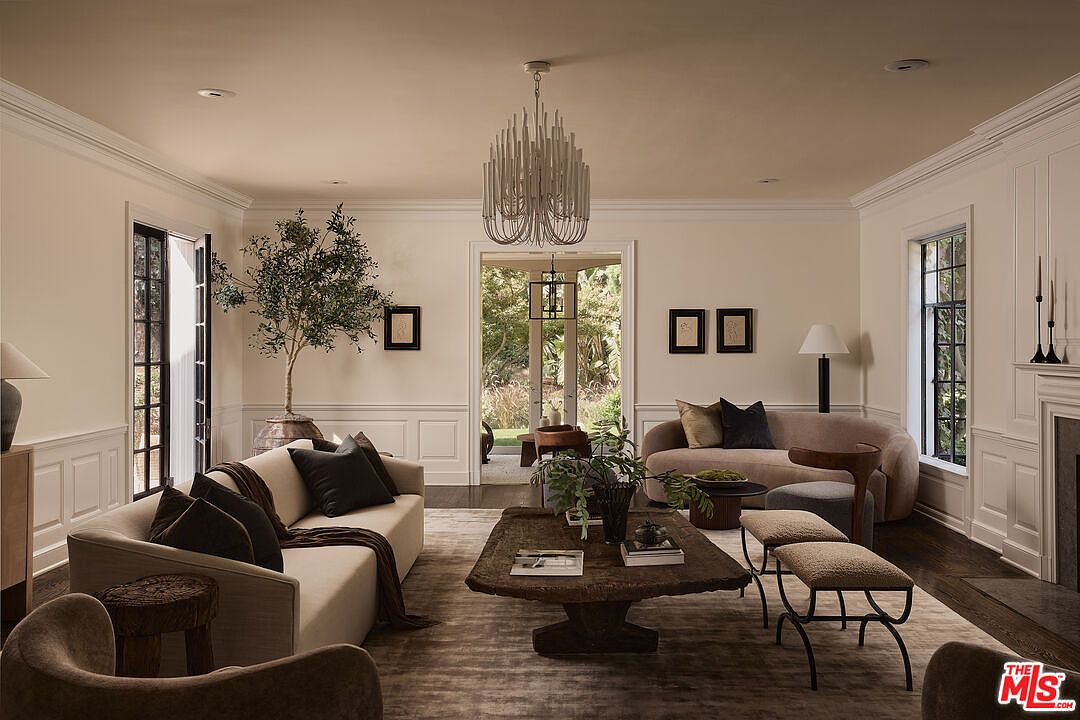 This is an interior shot of a living room featuring a neutral color palette with beige walls and dark wood floors. The room is furnished with two sofas, a unique wooden coffee table, and a modern chandelier. Natural light streams in through the windows, creating a warm and inviting atmosphere.