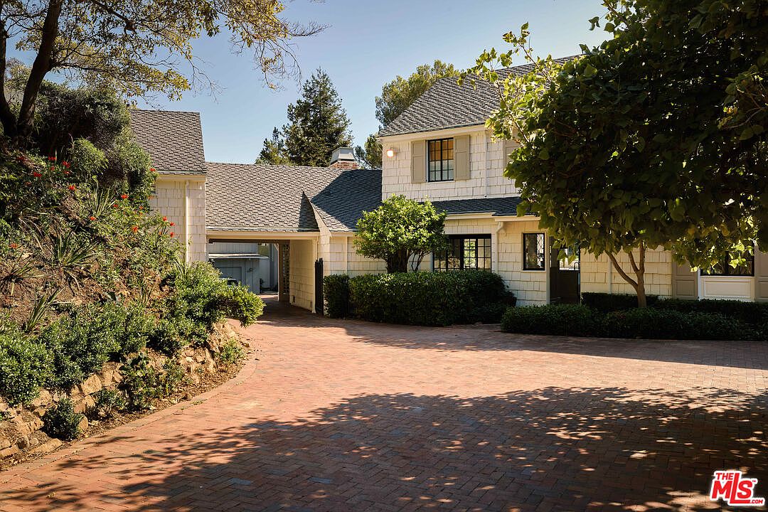 This is a front view of a charming two-story house with a brick driveway. The house features light-colored siding, dark-framed windows, and a gray shingle roof. Lush greenery surrounds the property, adding to its curb appeal and creating a welcoming atmosphere.