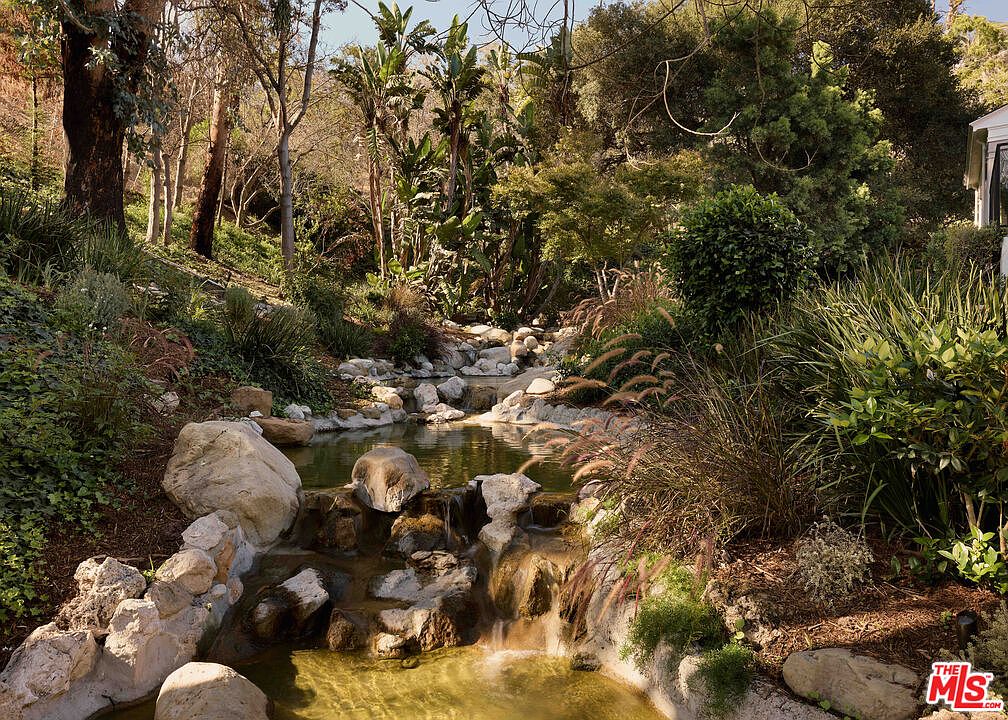 This image showcases a lush and serene garden featuring a rock-lined stream with small waterfalls and pools. The landscape is filled with a variety of plants, including trees, shrubs, and ornamental grasses, creating a tranquil and natural setting. The overall impression is one of a peaceful and well-maintained outdoor space, perfect for relaxation and enjoyment.