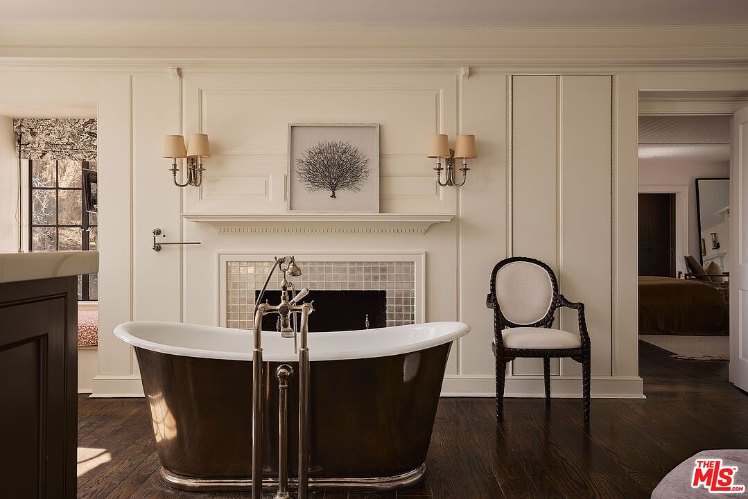 This is a luxurious primary bathroom featuring a freestanding, dark-colored bathtub with chrome fixtures as a focal point. The room is decorated with elegant wall paneling, sconce lighting, and a framed artwork above a faux fireplace. A chair sits to the side, and an open doorway leads to a bedroom, creating a sense of spaciousness and connectivity.