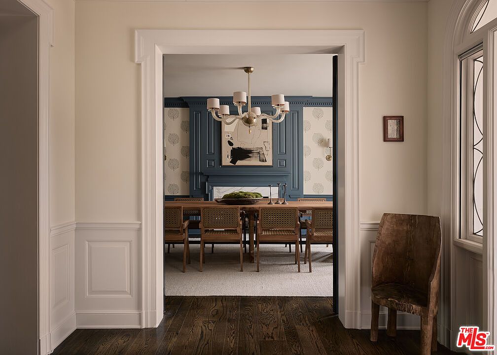 The image showcases a dining room viewed through a doorway, featuring a long wooden table with woven chairs set on a light-colored rug. The room is decorated with blue paneled walls and patterned wallpaper, complemented by a modern chandelier. A wooden chair sits in the foreground, adding a rustic touch to the elegant space.