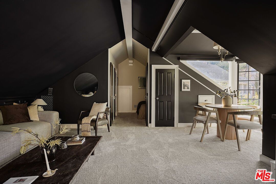 This interior shot showcases a stylish living room with a unique attic conversion. The room features dark-painted walls and ceiling, creating a cozy atmosphere, complemented by light-colored carpet and furniture. A round dining table with chairs sits near a window, while a sofa and coffee table occupy the other side of the room, creating a functional and aesthetically pleasing space.