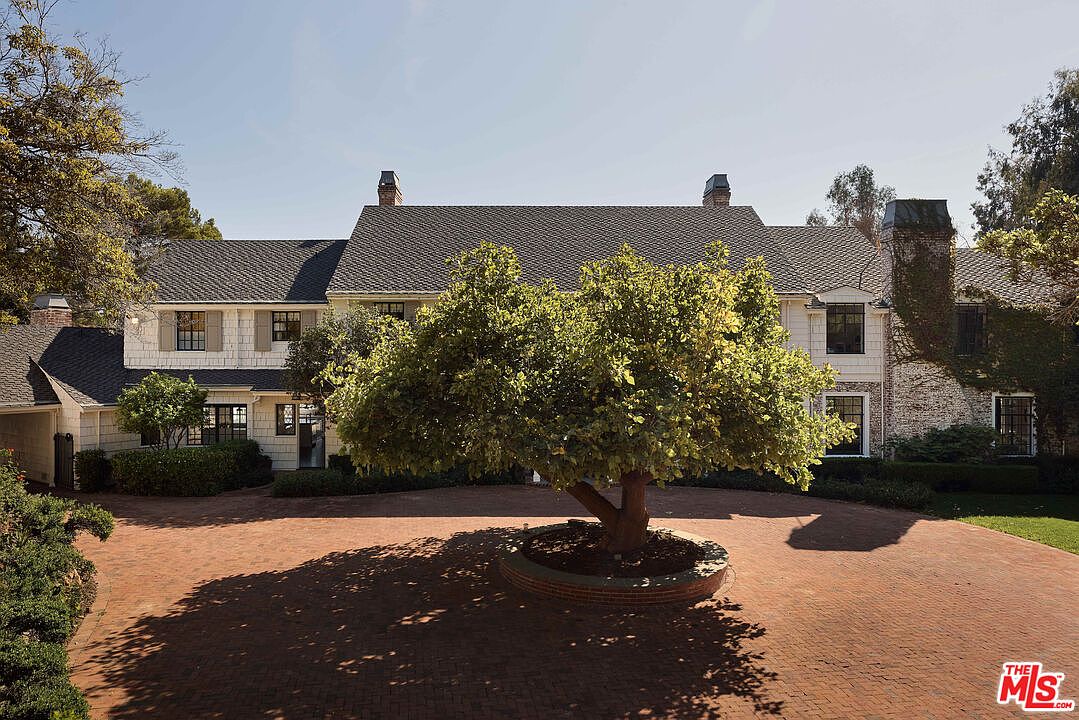 This is a front view of a large, traditional-style house with a brick driveway. The house features a gray roof, white siding, and multiple chimneys. A mature tree is centered in the driveway, adding to the property's curb appeal and providing shade.
