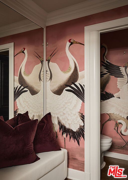 This image showcases a stylish guest bathroom featuring a unique crane-themed wallpaper in shades of pink and white. The room includes a toilet and a small seating area with plush burgundy pillows. The overall design creates a sophisticated and artistic ambiance, making it a memorable space for guests.